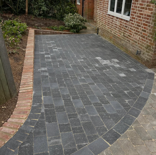 A social media post showing a curved driveway paved with dark gray rectangular stone slabs, bordered by red brick and green shrubs, with the post captioned for earthscapeconstruction.