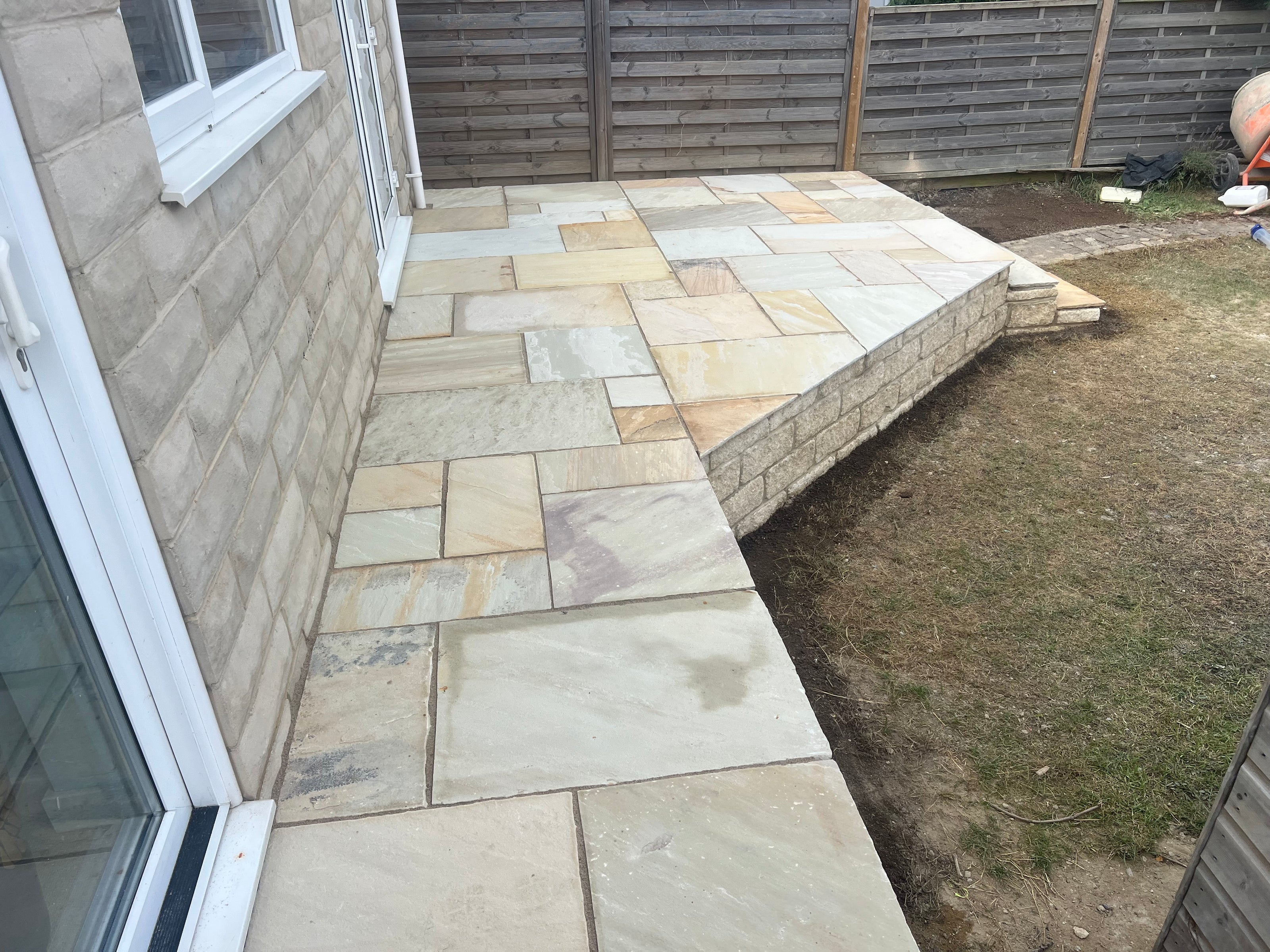 Paved patio area next to a house with grass on one side and retaining wall.